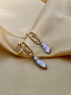 Close-up of a  luxury grey baroque keshi pearl drop earrings with a sculptural gold double-oval link stud. The image highlights the iridescent nacre and organic shape of the pearl, styled against a soft matte ivory background, representing high-end commercial jewelry photography