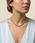 Close-up editorial portrait of a woman wearing a gold curb chain necklace with a central grey baroque pearl. The pearl has a silvery luster and sits on the collarbone. The model is wearing a deep V-neck light blue silk top against a warm ivory background