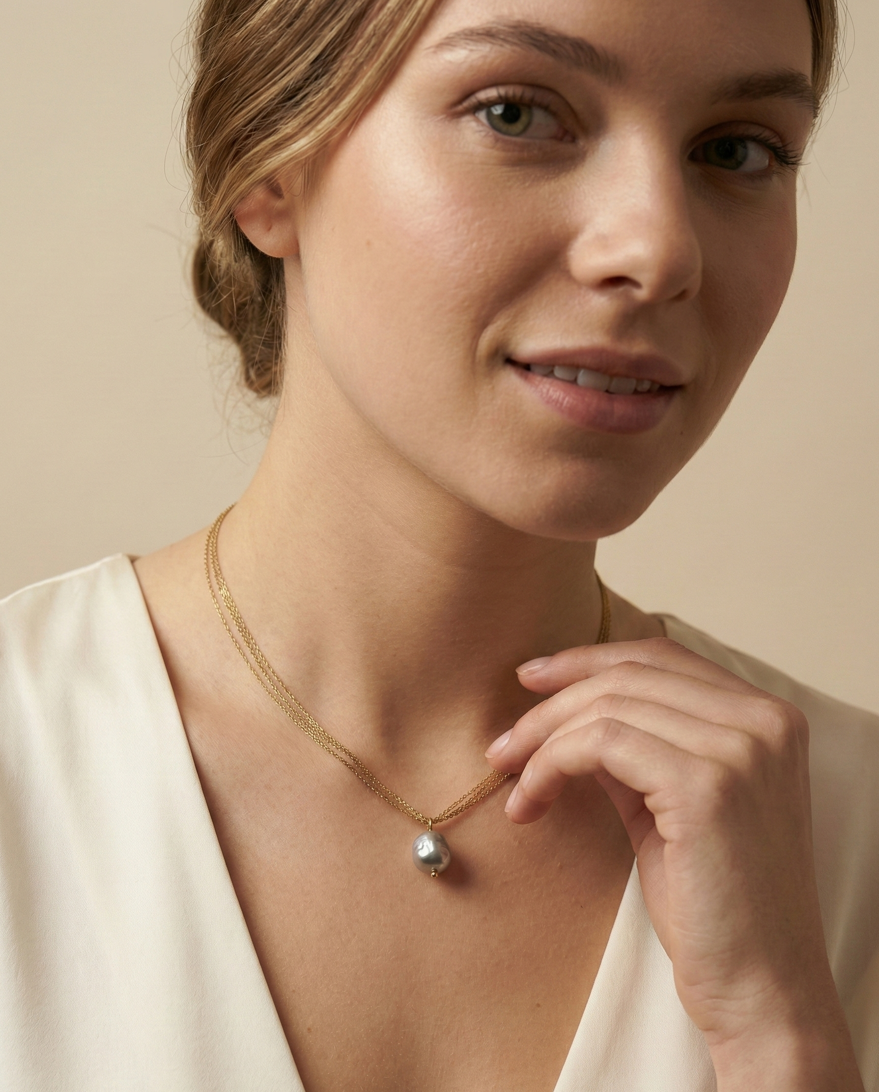 Close-up detail of a grey baroque pearl pendant with metallic luster, suspended on a delicate multi-strand gold chain. The necklace sits elegantly on a model's collarbone against an ivory silk V-neck top