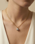Close-up detail of a grey baroque pearl pendant with metallic luster, suspended on a delicate multi-strand gold chain. The necklace sits elegantly on a model's collarbone against an ivory silk V-neck top