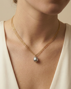 Close-up detail of a grey baroque pearl pendant with metallic luster, suspended on a delicate multi-strand gold chain. The necklace sits elegantly on a model's collarbone against an ivory silk V-neck top