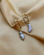 Close-up of a  luxury grey baroque keshi pearl drop earrings with a sculptural gold double-oval link stud. The image highlights the iridescent nacre and organic shape of the pearl, styled against a soft matte ivory background, representing high-end commercial jewelry photography