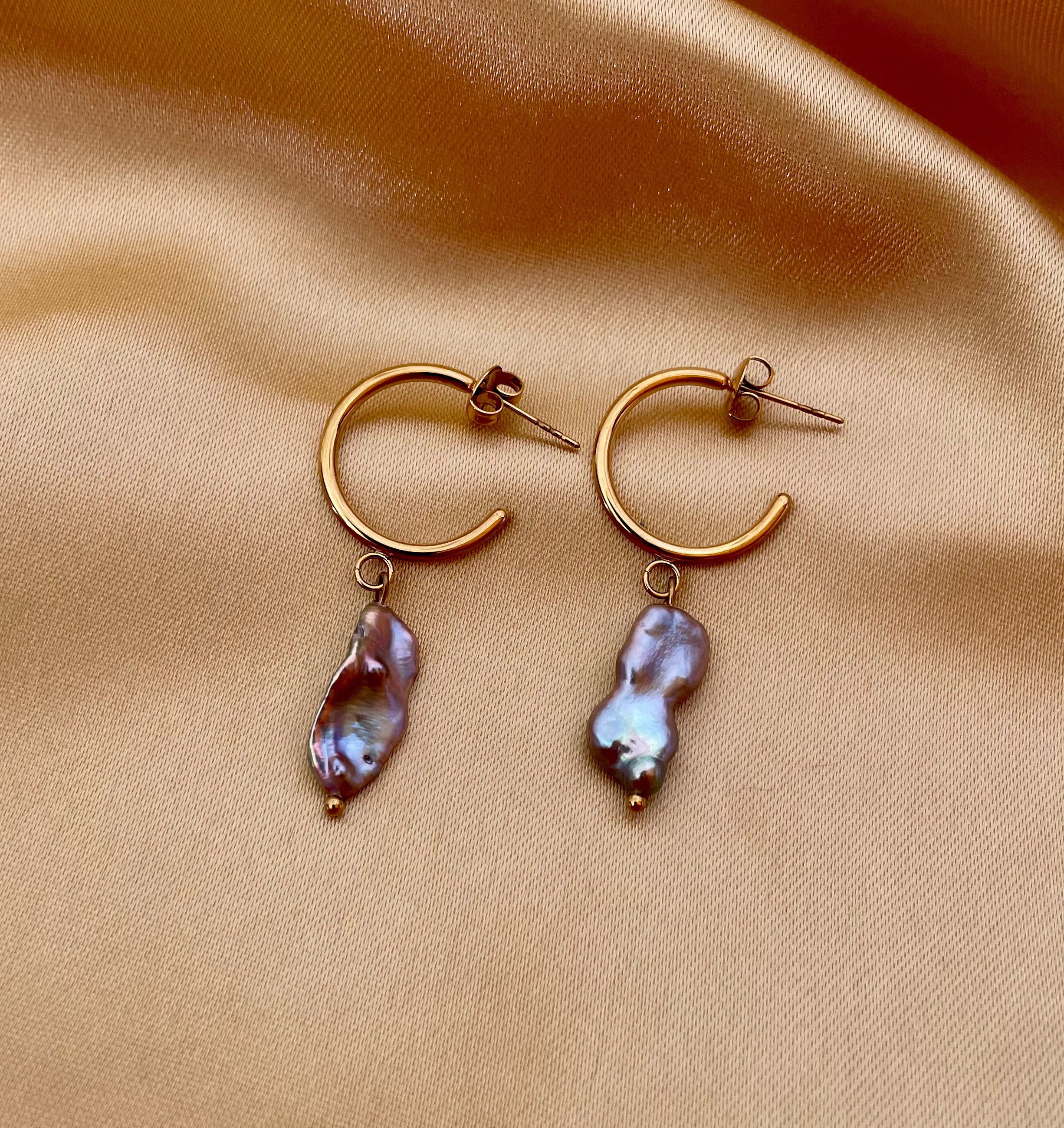 Close-up commercial jewelry photography of a gold huggie hoop earring featuring a lustrous grey keshi pearl drop. The organic, baroque-shaped pearl has high-quality nacre iridescence, realistic skin texture, and a warm, matte ivory background, highlighting the minimalist luxury aesthetic