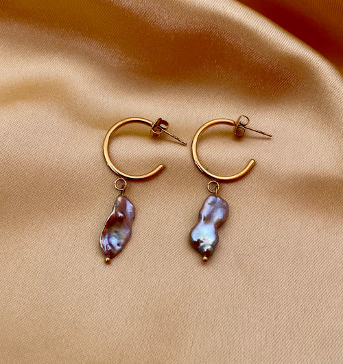 Close-up commercial jewelry photography of a gold huggie hoop earring featuring a lustrous grey keshi pearl drop. The organic, baroque-shaped pearl has high-quality nacre iridescence, realistic skin texture, and a warm, matte ivory background, highlighting the minimalist luxury aesthetic