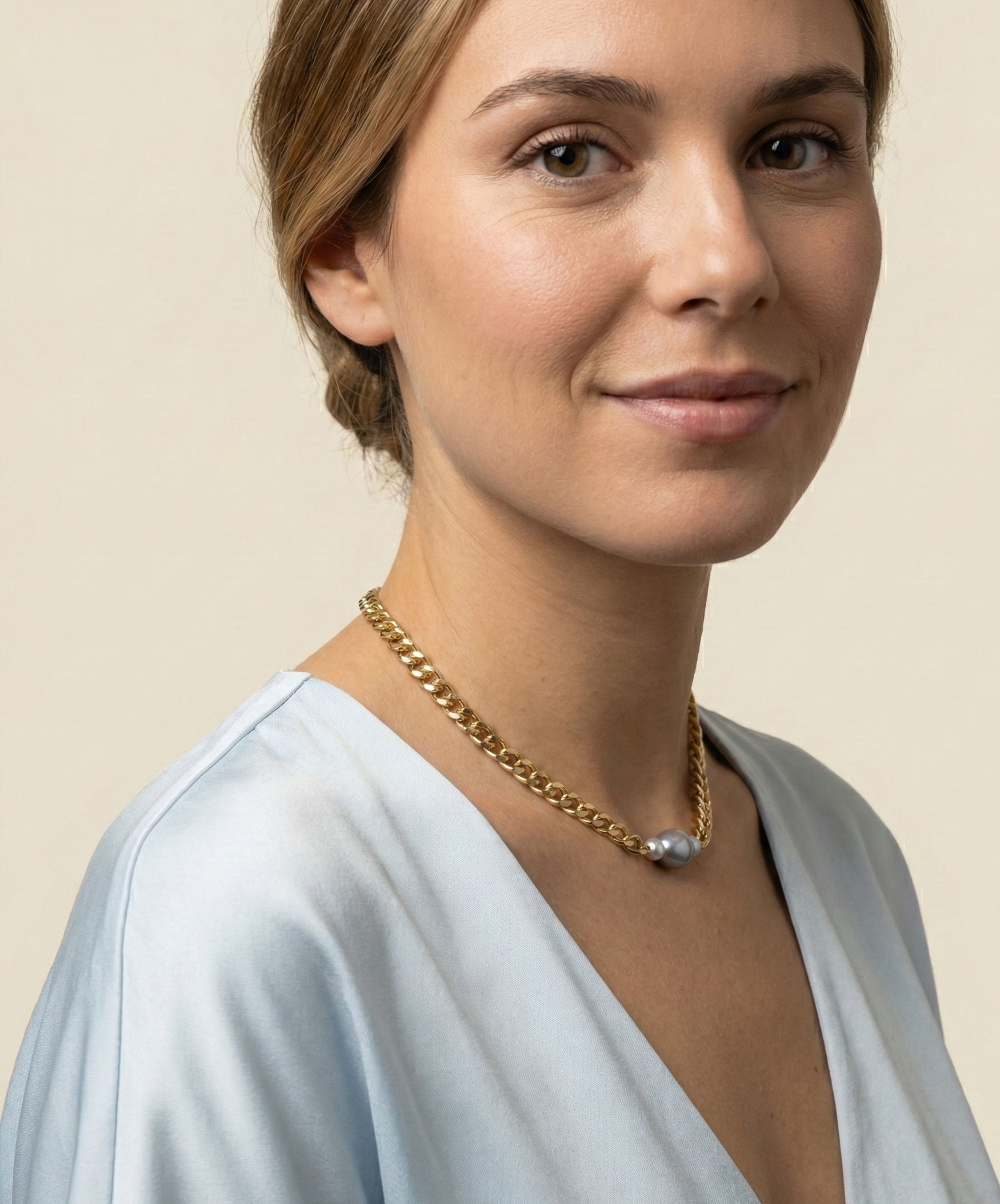 Model styling a grey baroque pearl pendant necklace on a chunky gold chain. The necklace is shown worn at collarbone length paired with a blue silk blouse, highlighting the pearl's iridescent shine