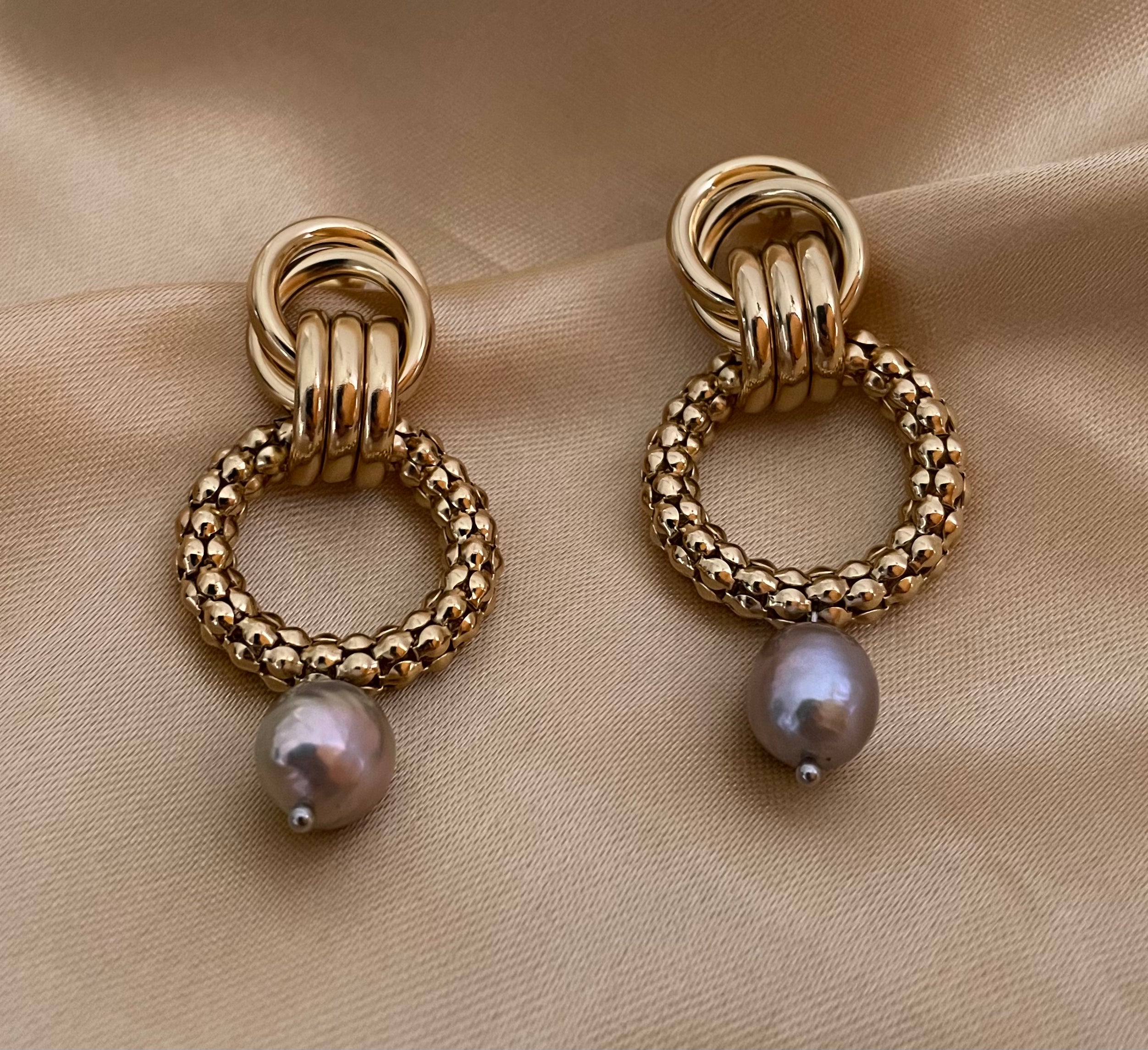 Close-up detail of textured gold interlocking hoop earrings featuring a lustrous metallic grey pearl drop. The design includes a beaded gold circle and polished gold links