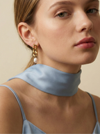 Editorial beauty portrait of a model wearing chunky gold link pearl earrings paired with a light blue silk scarf. The image highlights luxury jewelry trends and elegant styling