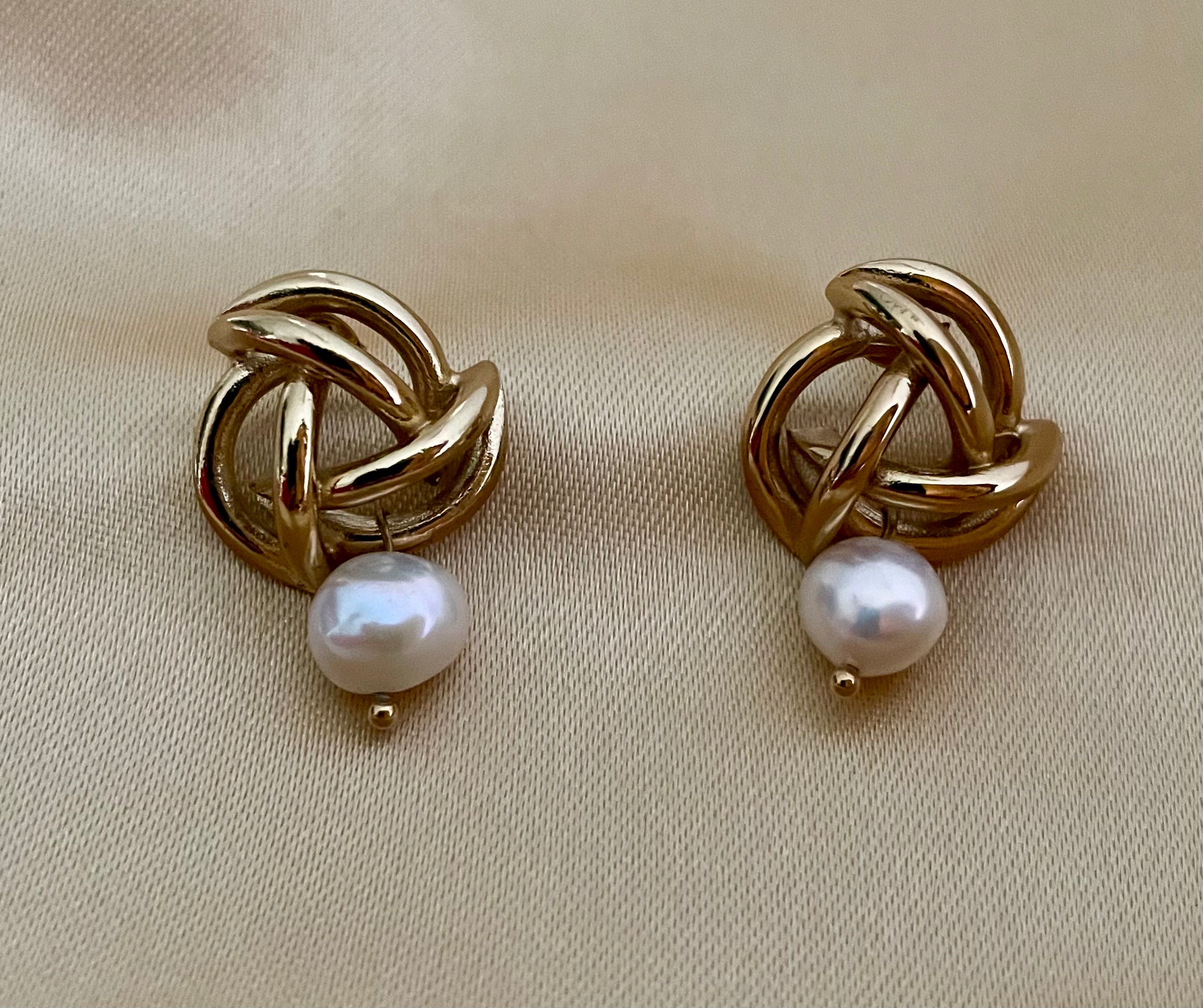 Close-up of gold twisted knot earrings with a white pearl drop, worn by a model in a cream silk blouse