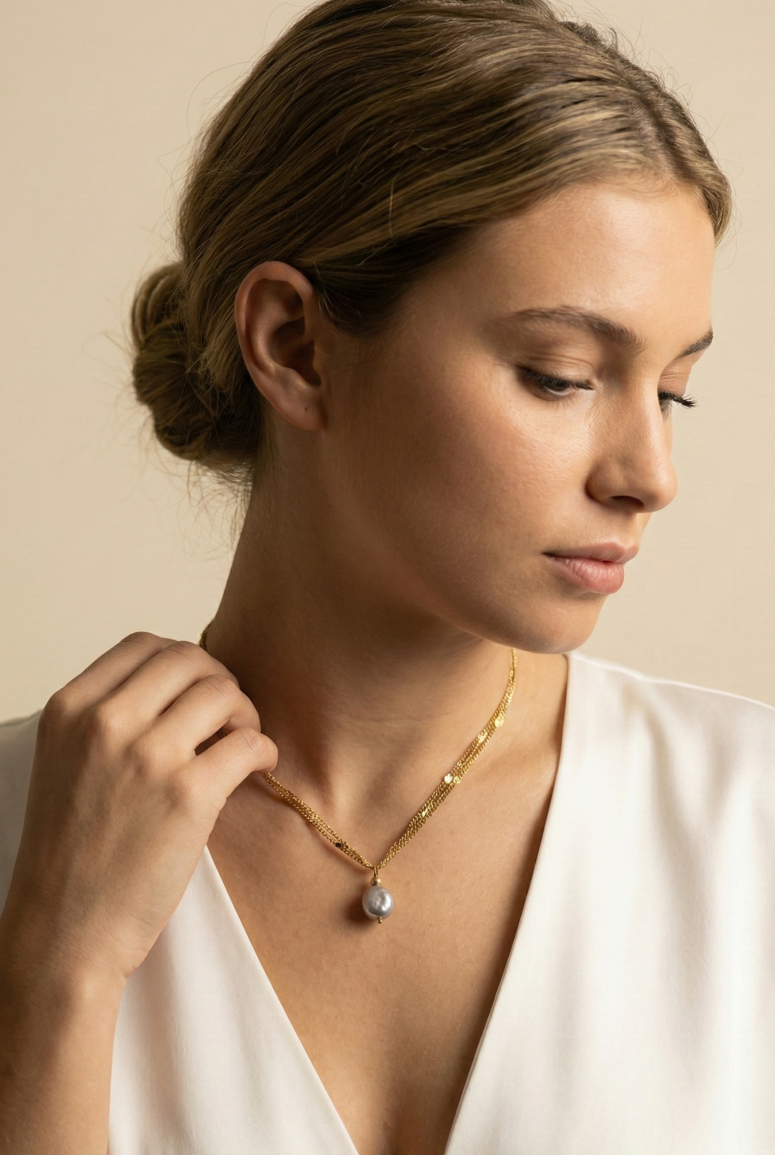 Close-up of a delicate triple-strand gold chain necklace featuring a single grey baroque pearl pendant, worn by a model in a white silk top
