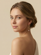 Side profile of a woman wearing contemporary gold U-shaped safety pin earrings with a dangling pink pearl. High-end commercial jewelry photography highlighting the pearl's luster against a matte ivory background