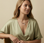 A young woman modeling a modern silver pearl necklace with a curved bar pendant, styled with a deep V-neck olive green silk top in soft studio lighting.