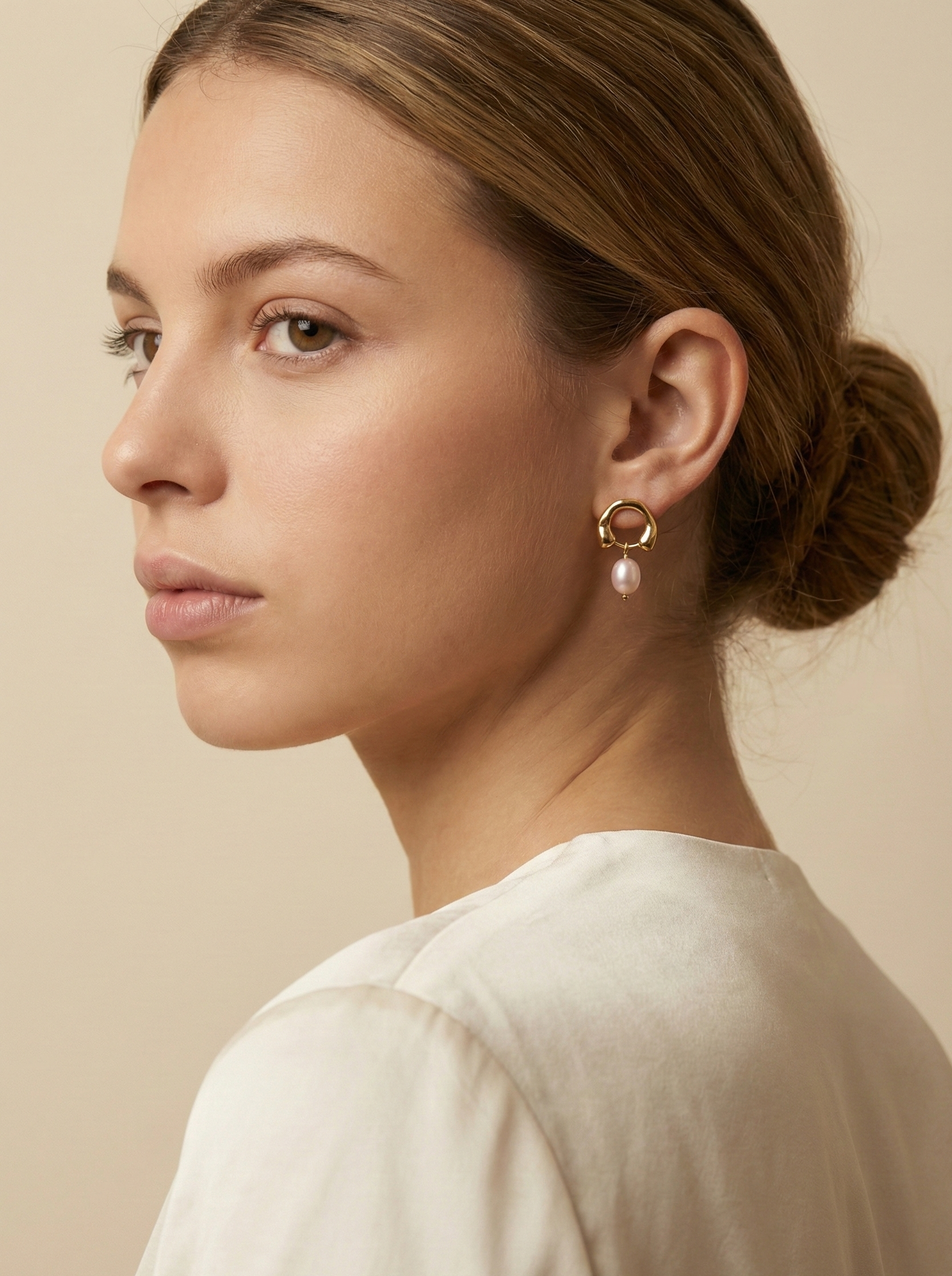 Close-up detail of organic gold huggie hoop earrings featuring a high-luster pink oval freshwater pearl drop, worn on a model's ear against a matte ivory background