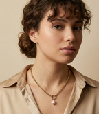A model with curly brown hair in a bun wearing a beige silk shirt and a Parisian chic gold chain necklace with a pink baroque pearl drop