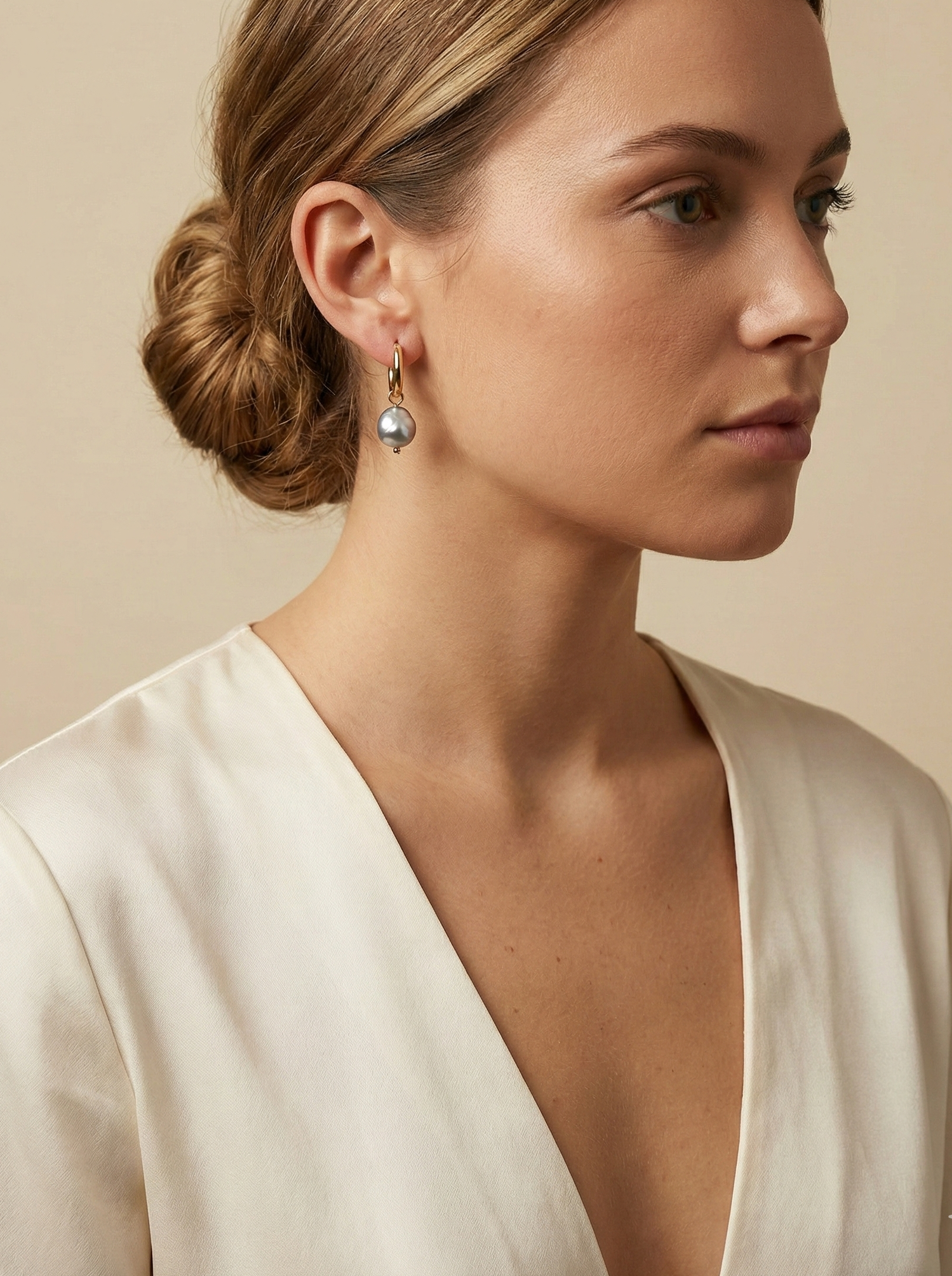 Editorial beauty portrait of a model with a low bun wearing unique grey pearl drop earrings and a deep V-neck cream silk shirt, styled in a Parisian chic aesthetic with warm lighting