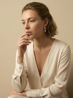 Editorial close-up of a model in a Parisian chic style wearing an ivory silk shirt, posing with her hand gently on her lips in a contemplative look. She is wearing unique, organic-shaped gold earrings featuring lustrous pink oval pearl drops.