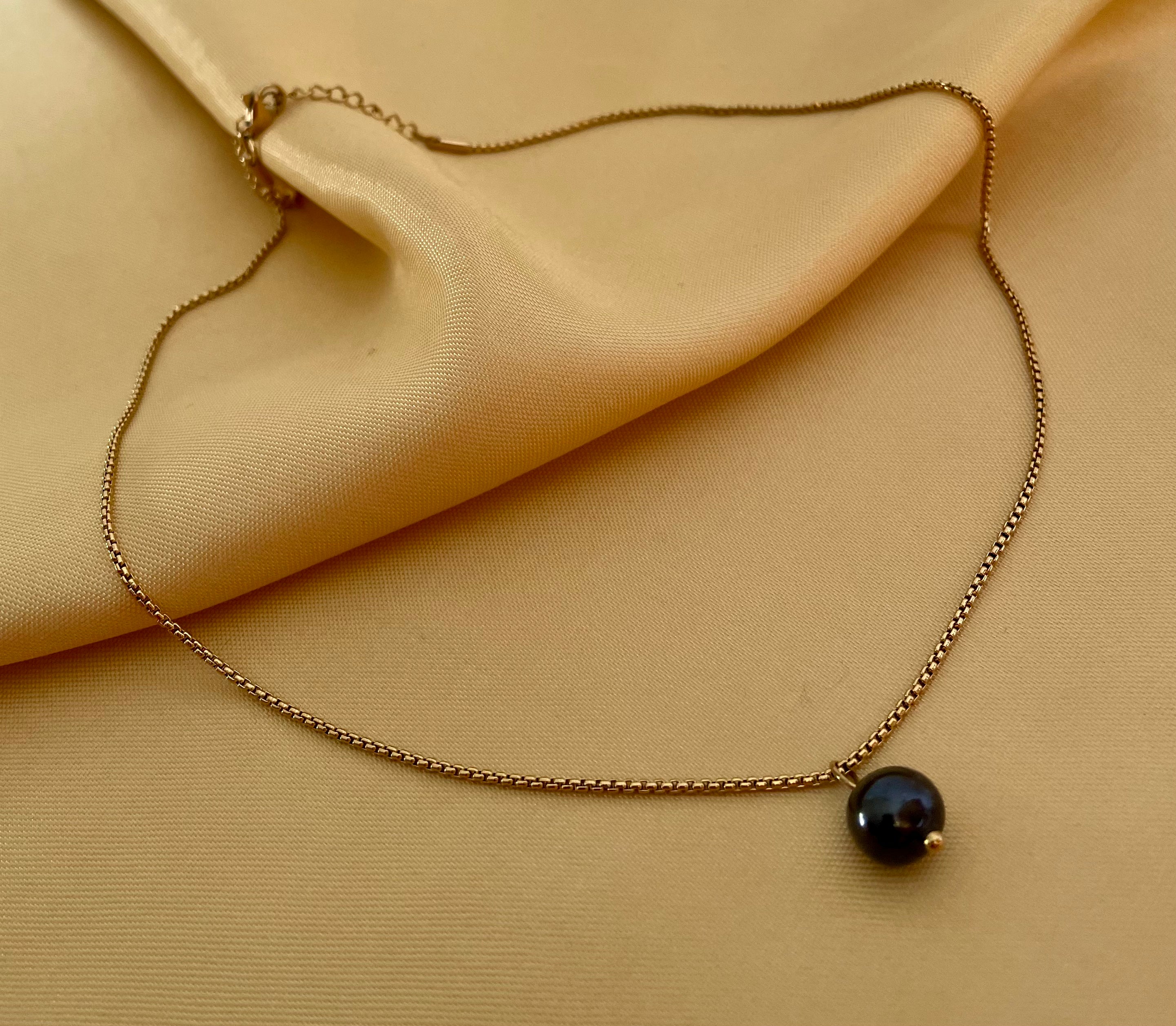 Close-up of a round black pearl solitaire pendant on a delicate gold box chain