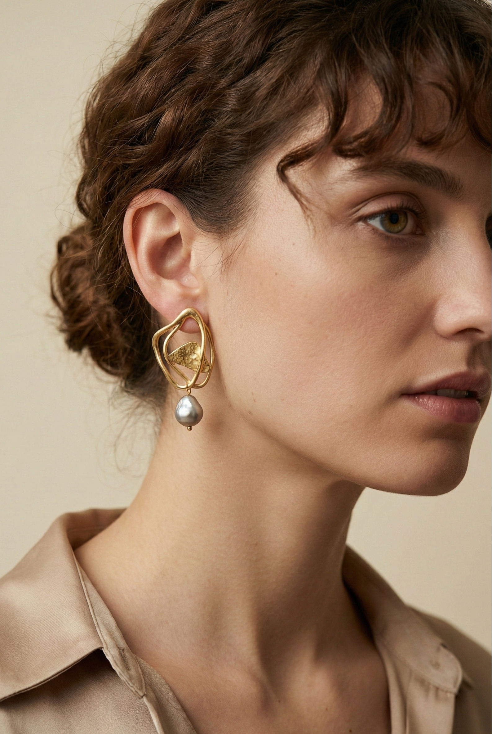 Close-up luxury jewelry editorial featuring sculptural gold open-frame earrings with a lustrous grey pearl drop. The model, with blonde curly hair, wears an ivory silk scarf and camisole, highlighting the textured gold metal and silver iridescent pearl against a soft warm studio background