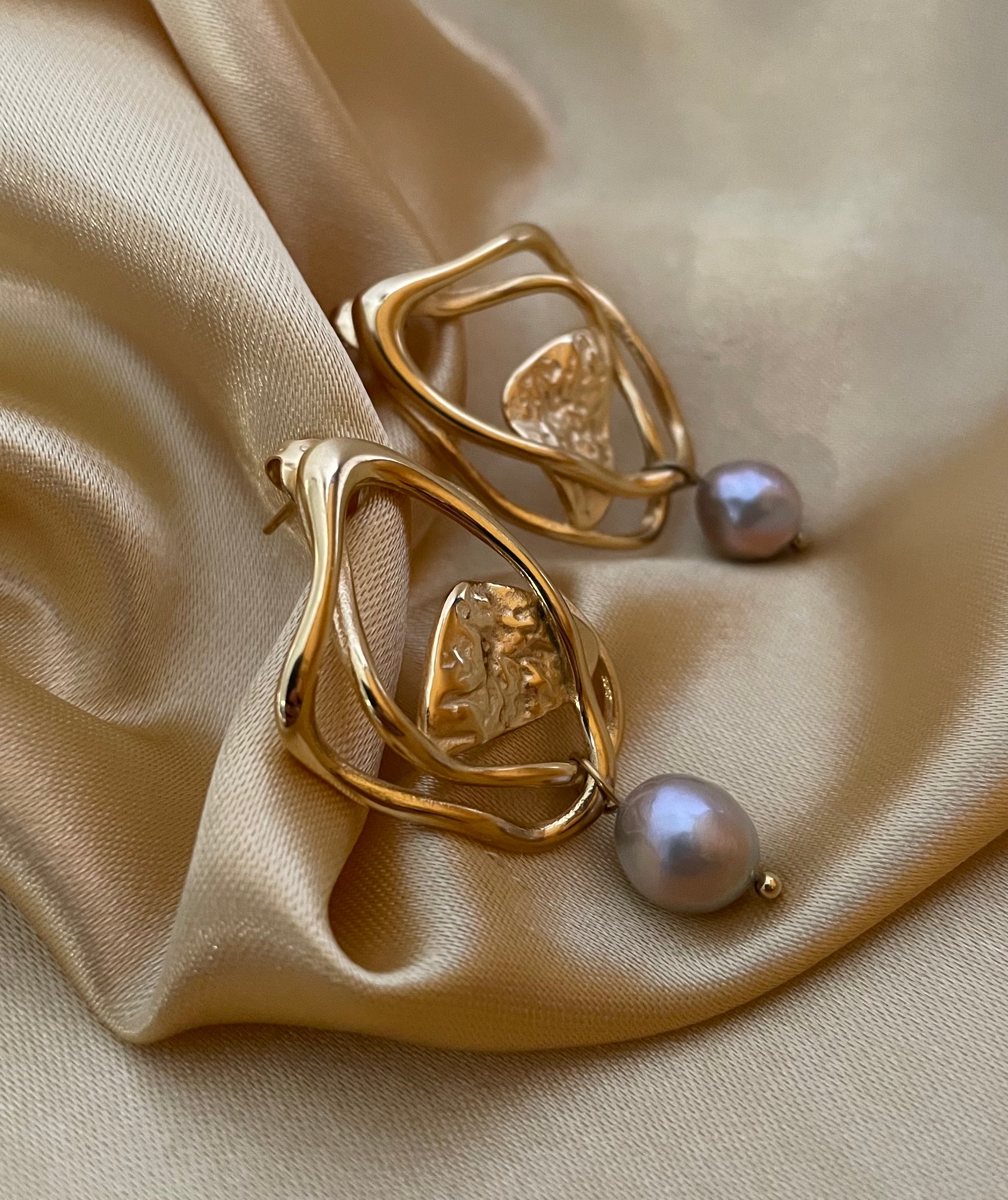 Close-up luxury jewelry editorial featuring sculptural gold open-frame earrings with a lustrous grey pearl drop