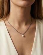 Close-up of a delicate silver curb chain necklace featuring a single white baroque pearl pendant with high luster, worn by a female model in an ivory silk top, showcasing the necklace length and elegant draping