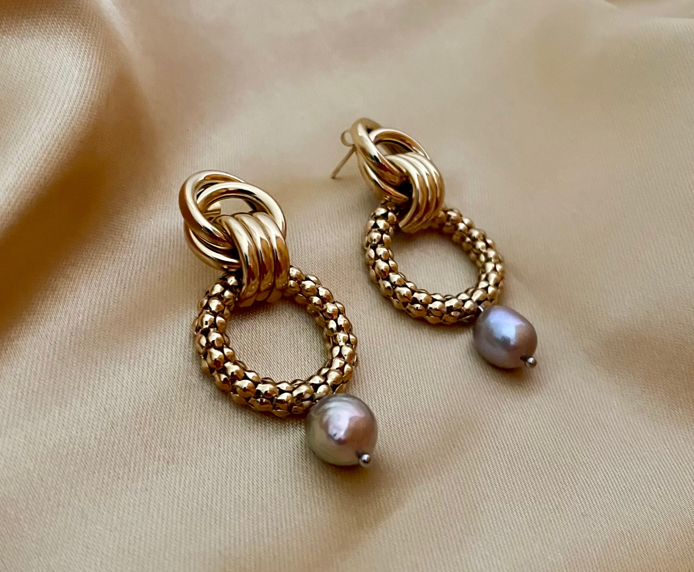 Close-up detail of textured gold interlocking hoop earrings featuring a lustrous metallic grey pearl drop. The design includes a beaded gold circle and polished gold links