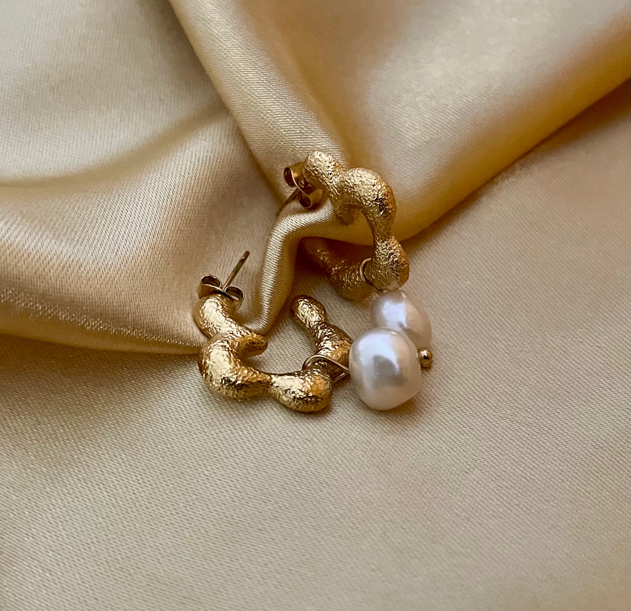 Close-up of textured gold huggie hoop earrings featuring a lustrous white pearl drop. The earrings have an organic, hammered gold finish
