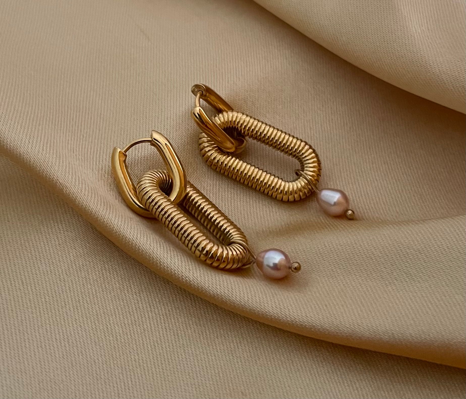 Close-up of textured gold oval link earrings featuring a dainty pink pearl drop. High-resolution commercial jewelry photography showcasing ribbed gold detailing and lustrous pearl against a soft matte ivory background.
