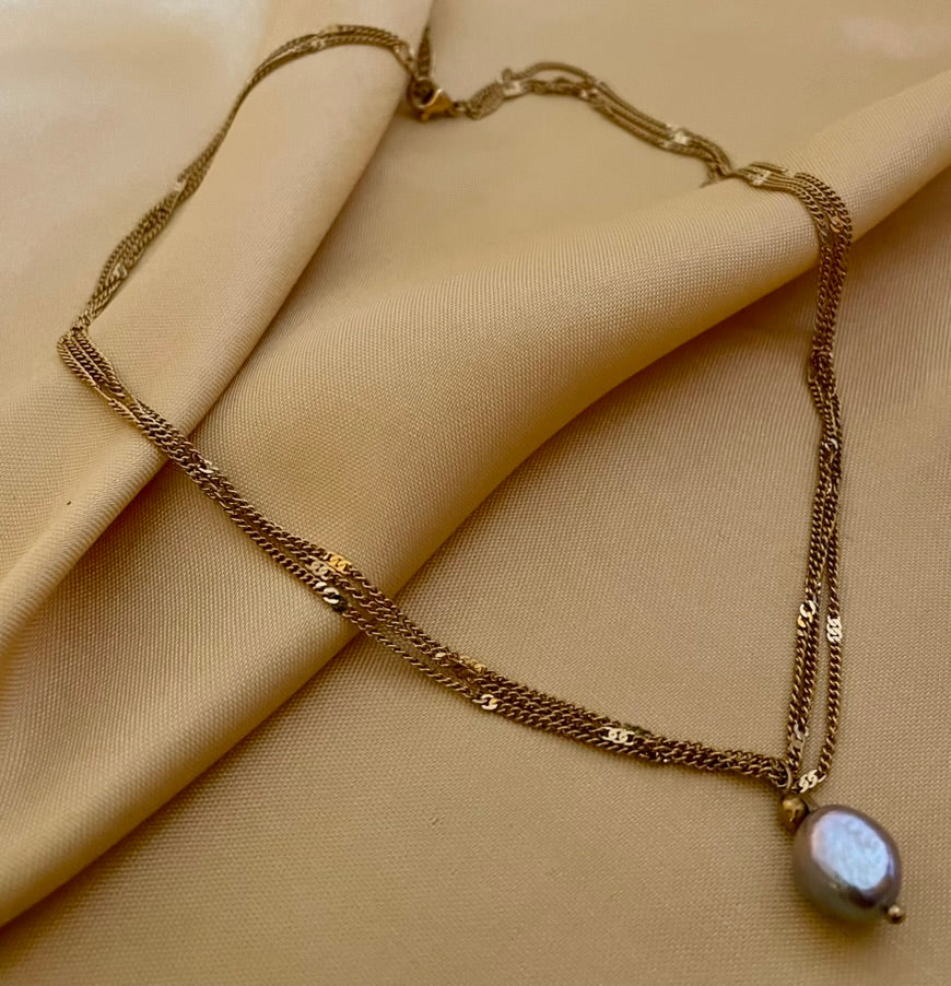 Close-up of a delicate triple-strand gold chain necklace featuring a single grey baroque pearl pendant, worn by a model in a white silk top.