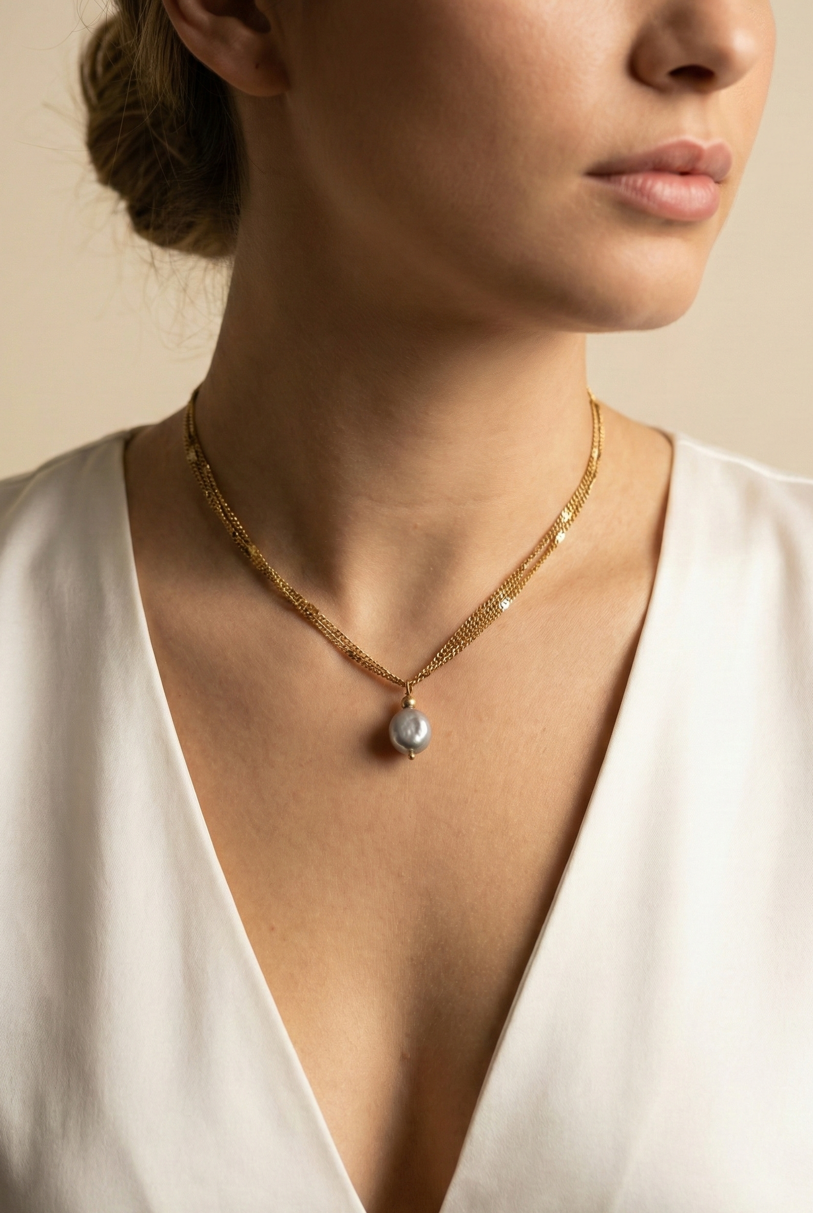 Close-up of a delicate triple-strand gold chain necklace featuring a single grey baroque pearl pendant, worn by a model in a white silk top