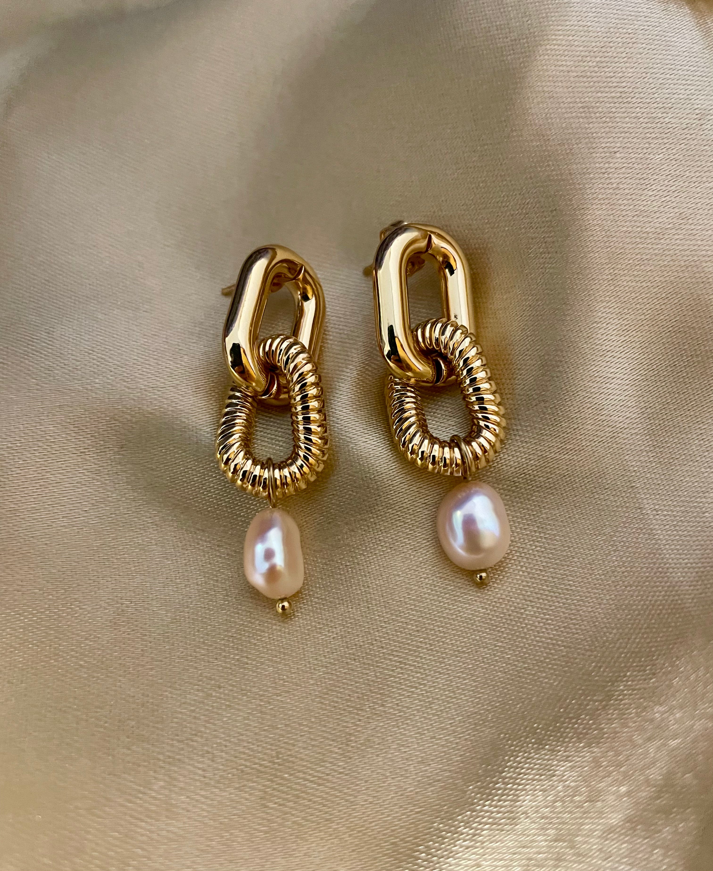Close-up of sculptural gold link earrings featuring a twisted rope texture and hanging freshwater baroque pearl drops. The earrings are displayed on a champagne silk fabric to highlight the pearl luster and high-shine gold finish