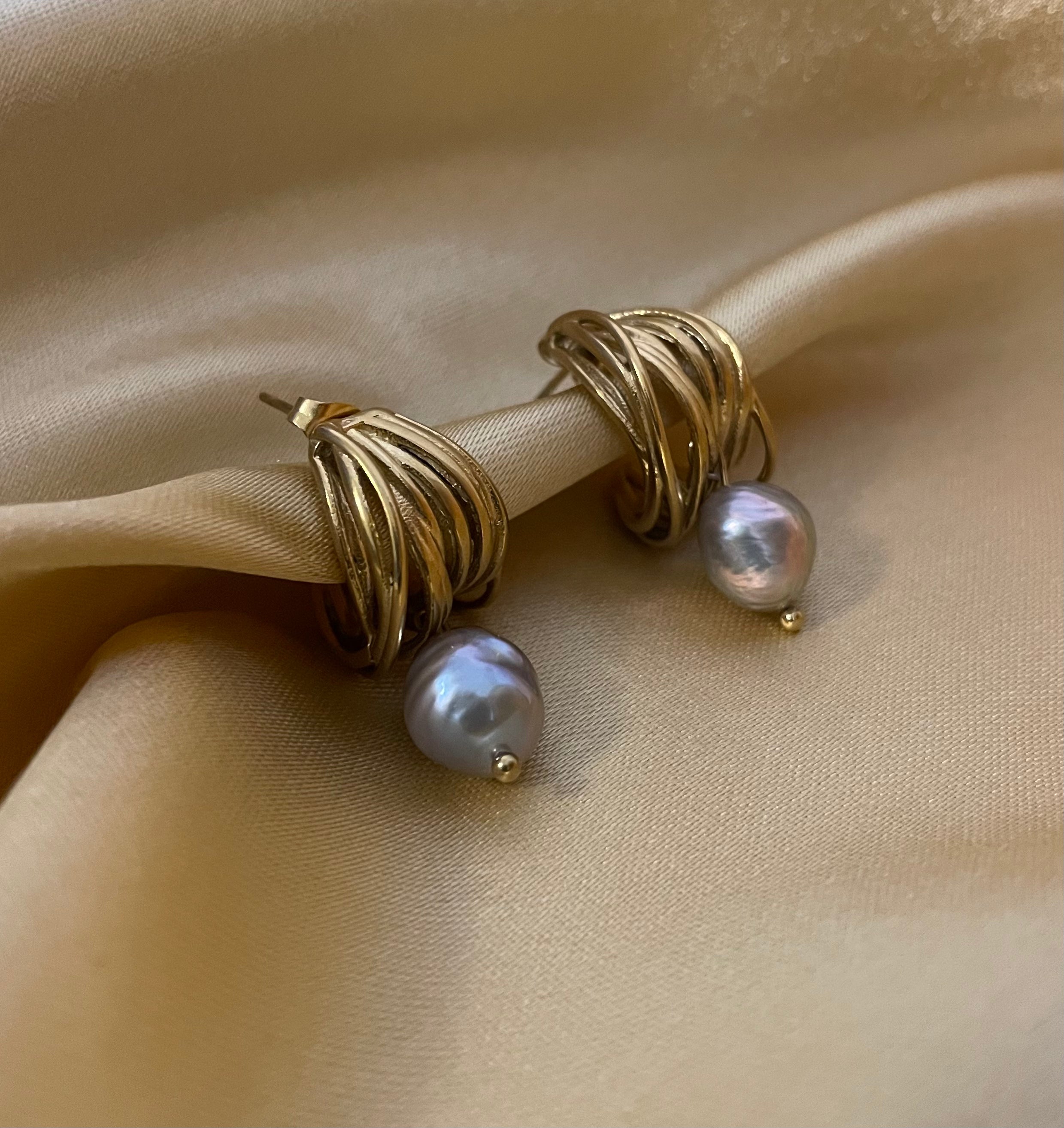 Luxury fashion jewelry photography of gold twist hoop earrings with dangling grey pearls. High-resolution close-up on the ear showing realistic skin texture, intricate gold detailing, and a sophisticated silver outfit.
